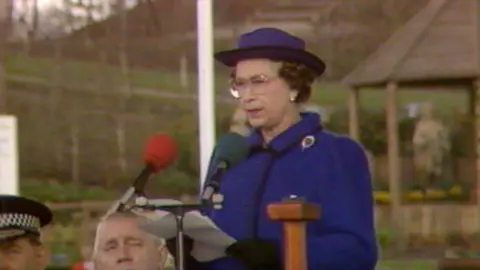 Queen Elizabeth II, a woman wearing glasses and a blue coat with a blue hat. She is standing at a lectern, holding a piece of paper and speaking into a red microphone. There are people sat behind her, including a police officer wearing a police cap. In the distance is an ornamental garden.