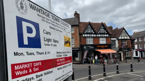 Wantage's Market Place. A small car park is pictured at the centre of a large town square. On the left is a parking information sign which gives information on parking times and costs. In the background are high street shops with a cafe outside featuring outside seating