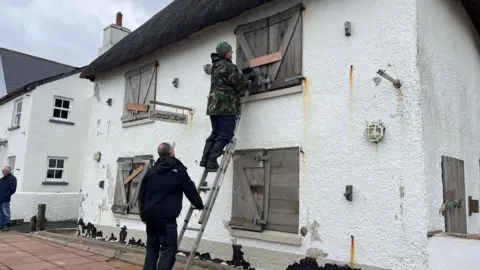 A man up a ladder putting flood defences on the windows or a white house on the Torcross seafront. There is a person holding the ladder at the bottom. 