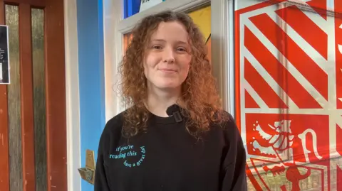 Beatrice has curly brown/ginger hair smiling into the camera. She is wearing a black jumper which has blue writing on the top left corner. Behind her is a tinted glass which is painted with a red crest.