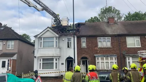 An end-terrace house that does not have a roof as it has been destroyed in a fire. Firefighters are standing in front of the property. 