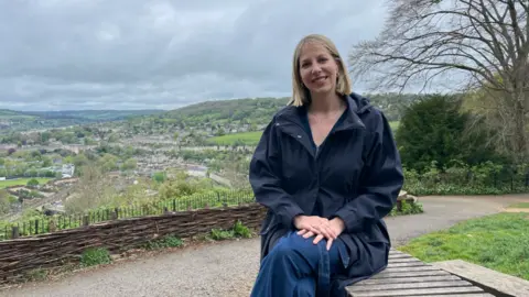 AWP A woman sits on a park bench with her hand resting on top of her crossed legs. She is sitting alongside a park which overlooks Bath, with rooftops, fields and trees visible in the background. It is an overcast day with grey clouds filling the sky.