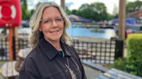 A woman with wavy blonde hair and glasses, wearing a black jacket with a leopard print top underneath, is pictured from the shoulders up, smiling at the camera.