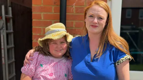 Sofia Akin A woman and young girl look into the camera smiling. The woman has long red hair and is wearing a blue shirt. She has her arm around her daughter, who is wearing a pink dress and yellow hat.