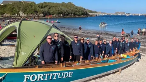 A group of people stand on a sandy beach beside a large blue wooden boat named 'FORTITUDE' with white lettering. A green tent is partially covering the boat. In the background, more people, boats, and water activities are visible in a bay or harbour, with vegetation and buildings along the distant shoreline.