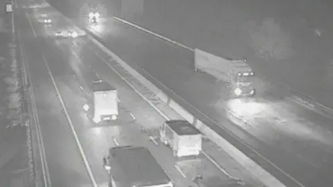 A traffic camera image of the M1 motorway at night time. Vehicles pass on each side of the road with their headlights on.