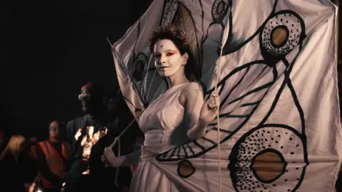 DAN B The May Queen wearing all white and white face paint with a large white banner behind her.