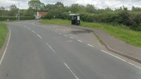 google A rural road bordered by hedges with a bus stop and layby on the righthand side.