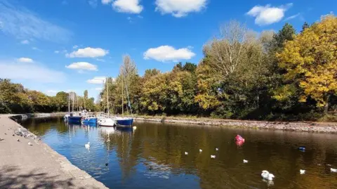 Geograph/Stephen Craven A large water feature with boats and swans. Concrete footpaths surround it.