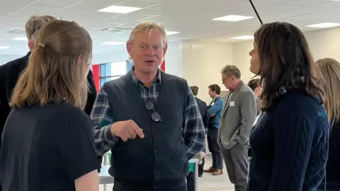 The actor Martin Clunes is speaking to a group of people including two women, whose backs are to the camera. Mr Clunes, wearing a blue check shirt and a dark blue gilet, is gesticulating with his right hand. Various people in suits can be seen behind them. 