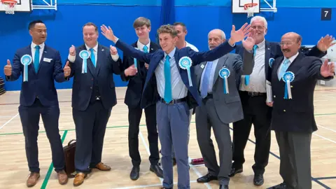 Reform UK candidates in the election count venue in Coalville, Leicestershire. They all sport blue rosettes and wear suits over white shirts and light blue ties.