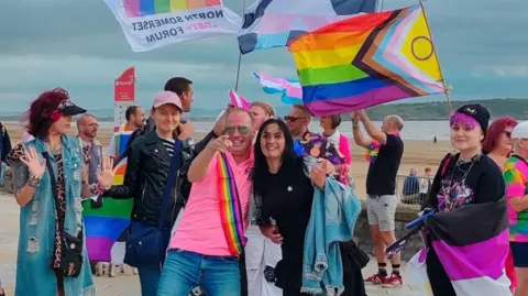 A group of people dressed in different colours and waving LGTBQ flags are standing by the beach in Weston-super-Mare. Several people are smiling towards the camera. 