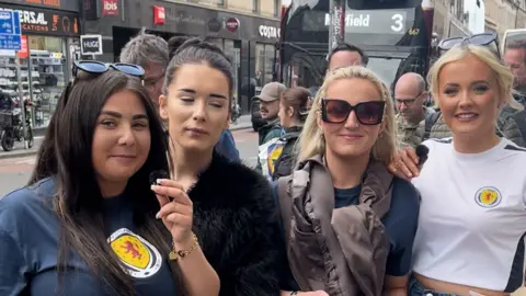 Four smiling women wear Scotland tops in an Edinburgh street
