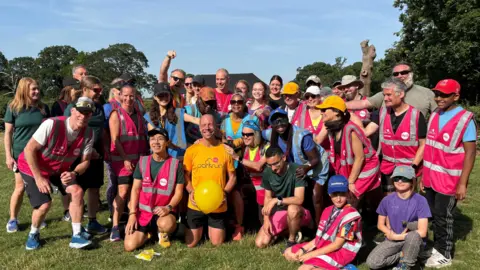 PA Media Wood wearing a yellow T-shirt with the word 'parkrun' printed across the front, holding a yellow balloon, surrounded by about two dozen people, most of whom are wearing sun hats, sunglasses and high visibility vests and smiling into the camera.