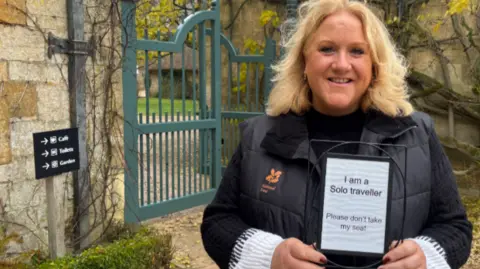 Sue is outside the gates to Hidcote Gardens in a midshot, smiling to camera and holding a sign saying 'I am a Solo traveller, please don't take my seat!'. She is in a black gilet with the national trust logo on it. There is a board behind her directing visitors to the cafe, toilets and garden.