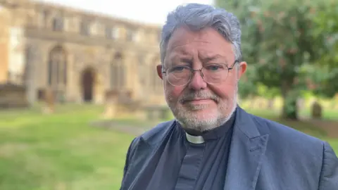 A man with grey hair and stubble, wearing glasses, a black suit and a clerical collar. He's looking at something off-camera, and standing in a graveyard with a church behind him.