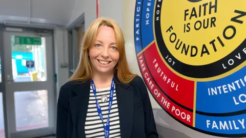 Kelly Davis stands in one of the school corridors next to a large circular sign that says "Faith is our foundation" in the centre. She is wearing a black and white striped t-shirt and a black blazer, with a necklace blue staff lanyard around her neck.