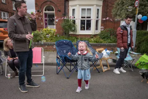 Getty Images A young girl shouts as she chases bubbles along a road as her father blows bubbles, outside houses at a street party in Middlesborough.