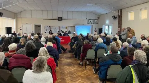 An audience of people sitting on chairs inside a hall as they listen into a public consultation pertaining to fire station closures in Dorset and Wiltshire