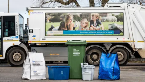 White waste bin lorry with green wheeler bin and other recycling containers in front of it including a white bag, a blue bag, a blue box, a grey caddy and a smaller grey caddy.