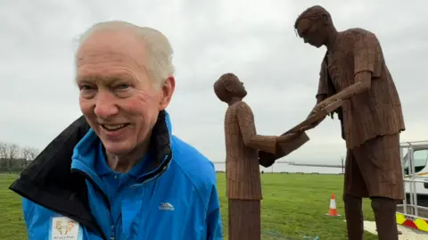 Brian Burnie, wearing a blue coat which says Daft as a Brush, stands in front of the bronze-coloured metal artwork which has been unveiled in South Shields. The artwork shows a young boy receiving a banjo from Len Gibson, a prisoner of war.