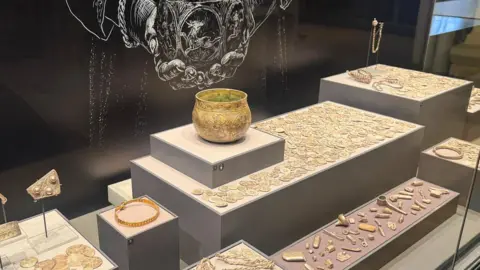 BBC/ Emily Johnson A silver bowl, coins and silver trinkets are displayed in a glass cabinet.