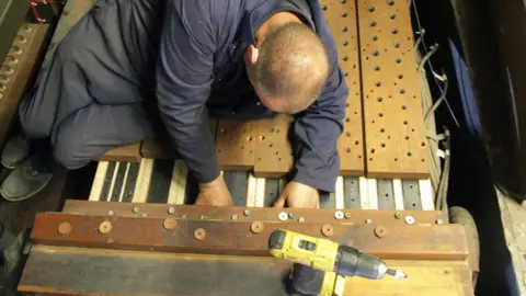 Duncan Smith Image of a man in blue work overalls, leaning over a section of an organ with a yellow drill in the foreground