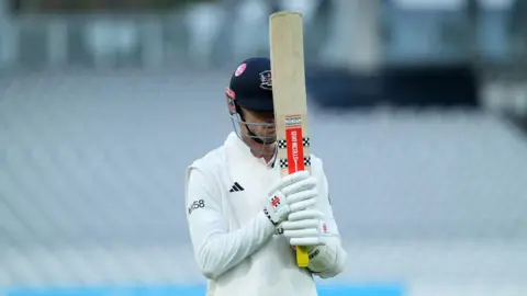 Ollie Price of Gloucestershire walks off the pitch with his bat held up towards the sky covering his face