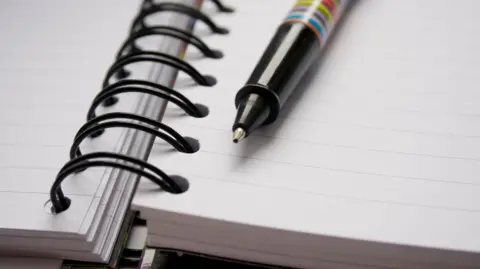 Close-up of an open spiral-bound notebook with lined pages. A diagonally placed pen featuring a colourful striped design rests on the notebook.