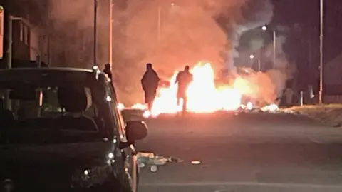 The silhouettes of people shrouded in darkness are seen in front of a large fire in a street lined with houses and a car in the foreground.