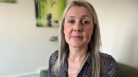 Emma Baugh/BBC News Mandy Geraghty, with blonde hair past her shoulder, has mascara and pink lipstick on. She is smiling and wearing a patterned top, and the background is blurred