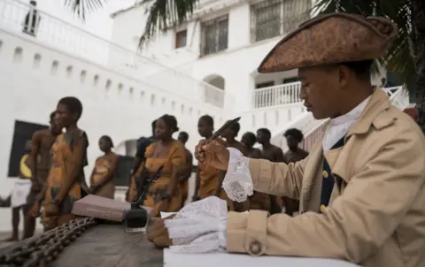 Ernest Ankomah / Getty Images A person in the foreground wears pre- Victorian-era clothing and signs a document with a quil while wearing a hat, as people dressed in scantily clad slave robes walk through the street in chains in the background, during a re-enactment of the trans-Atlantic slave trade in the courtyard of Christiansborg Castle in Accra, Ghana on 19 June.