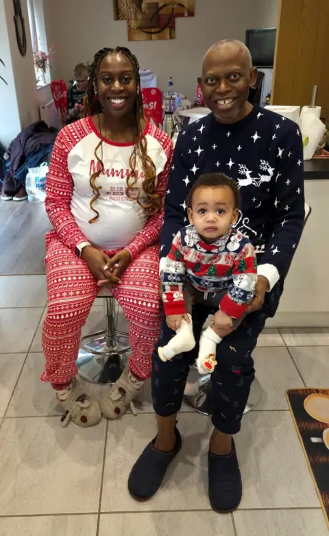 Kadilobari Peake A family portrait of a man sitting in a chair with his grandson on his lap and his daughter sitting next to him. They are wearing Christmas clothes. The man has cropped grey hair, his daughter has long braider hair and his grandson has short brown hair.