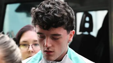 Layton Randle being taken into court. He has short brown curly hair and is wearing a white top with a green stripe and has a diamond earring. He is leaving a van and is being led by a security officer. 