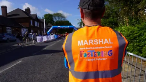 North East Lincolnshire Council A race marshal wearing an orange high-visibility vest stands by a road closure barrier with runners and spectators on the road in the background