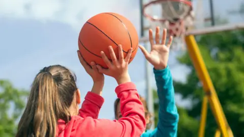 A young person seen from the back holding a basketball in the air as they get ready to take a shot. In the background there is a basketball hoops and another person standing in front of the first, holding their hand in the air.