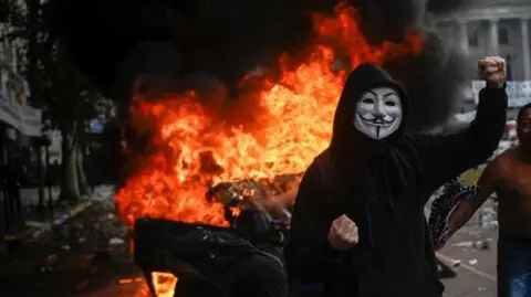 Reuters A person wearing a mask and a hoodie stands with his fist clenched in the air in front of a fire during protests turned violent in Buenos Aires, Argentina