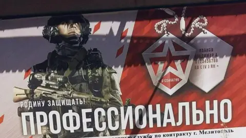 BBC Um pôster em vermelho e branco mostra um soldado russo segurando uma arma e usando equipamento de combate pronto para a batalha