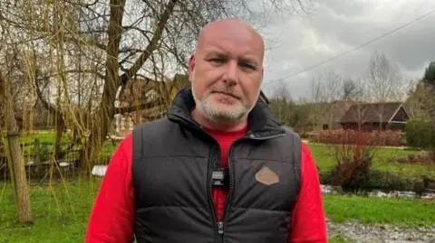 James Devine is pictured wearing a black body warmer over a long-sleeved red shirt. Behind him are trees, patches of grass and a waterway. 