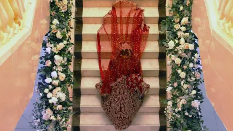 Maariya Kahn Maariya Kahn, who is wearing an embellished red and gold wedding dress with a veil, is walking down the staircase which is decorated with pink flowers.