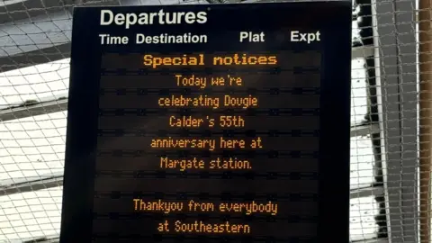 Southeastern A black digital train notice board. In orange writing it reads 'Special notices. Today we're celebrating Dougie Calder's 55th anniversary here at Margate Station. Thankyou from everybody at Southeastern.' 