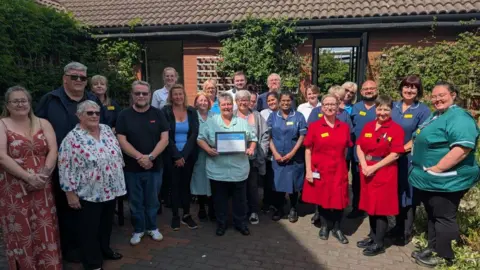 George Eliot Hospital NHS Trust Lisa Wheelhouse has short grey hair and is wearing a light blue shirt and black trousers. She is holding a framed award in her hands. Standing around her are a crowd of people, most of whom are wearing hospital uniform in dark blue, teal or red.
