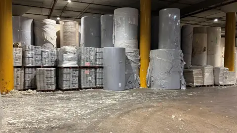 The inside of a factory, showing about a dozen large blue rolls, next to pallets of smaller blue rolls. There are some bright yellow pillars, and paper residue on the floor.