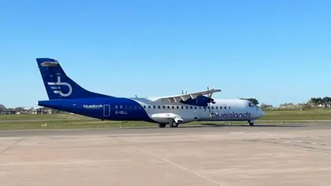 BBC A Blue Islands blue and white plane on a cream runway, with grass and blue sky behind.