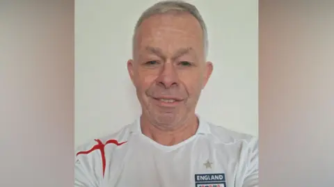 John Little John Little. He has short grey hair and blue eyes. He is wearing an England shirt and smiling at the camera.