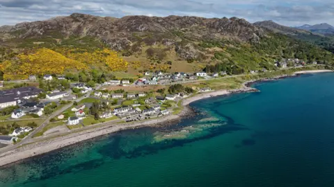 An aerial view of Gairloch. There are white-walled houses along a strip of shore between calm, green waters of the sea and a low range of rocky hills.
