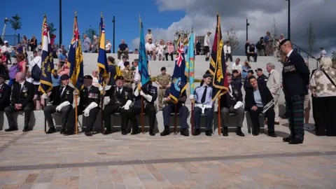 Members of the armed forces are sitting at the amphitheatre, holding flags. They are wearing uniform and havesome of the have medals.
