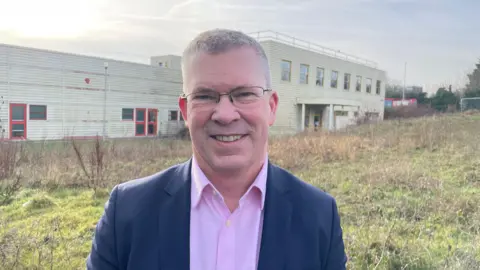 Laura Foster/BBC A man in a suit stands in front of an older, metal‑clad building surrounded by overgrown grass, with the structure’s faded red window frames and worn exterior visible in the background.