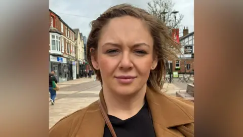 Elizabeth has brown hair. She is wearing a black top and a brown jacket and is standing in Ormskirk town centre looking at the camera.