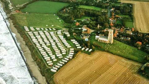 Mike Page An aerial image of a coastline, beach and village, showing a large field and a caravan park directly adjoining cliffs and beyond that a church, graveyard and housing 